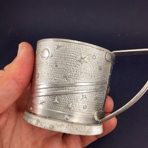 Peut inclure: Une tasse en m&eacute;tal argent&eacute; avec une poign&eacute;e, pr&eacute;sentant un motif sur le th&egrave;me de l'espace avec des &eacute;toiles et des plan&egrave;tes. La tasse a une surface textur&eacute;e et un fond c&ocirc;tel&eacute;.