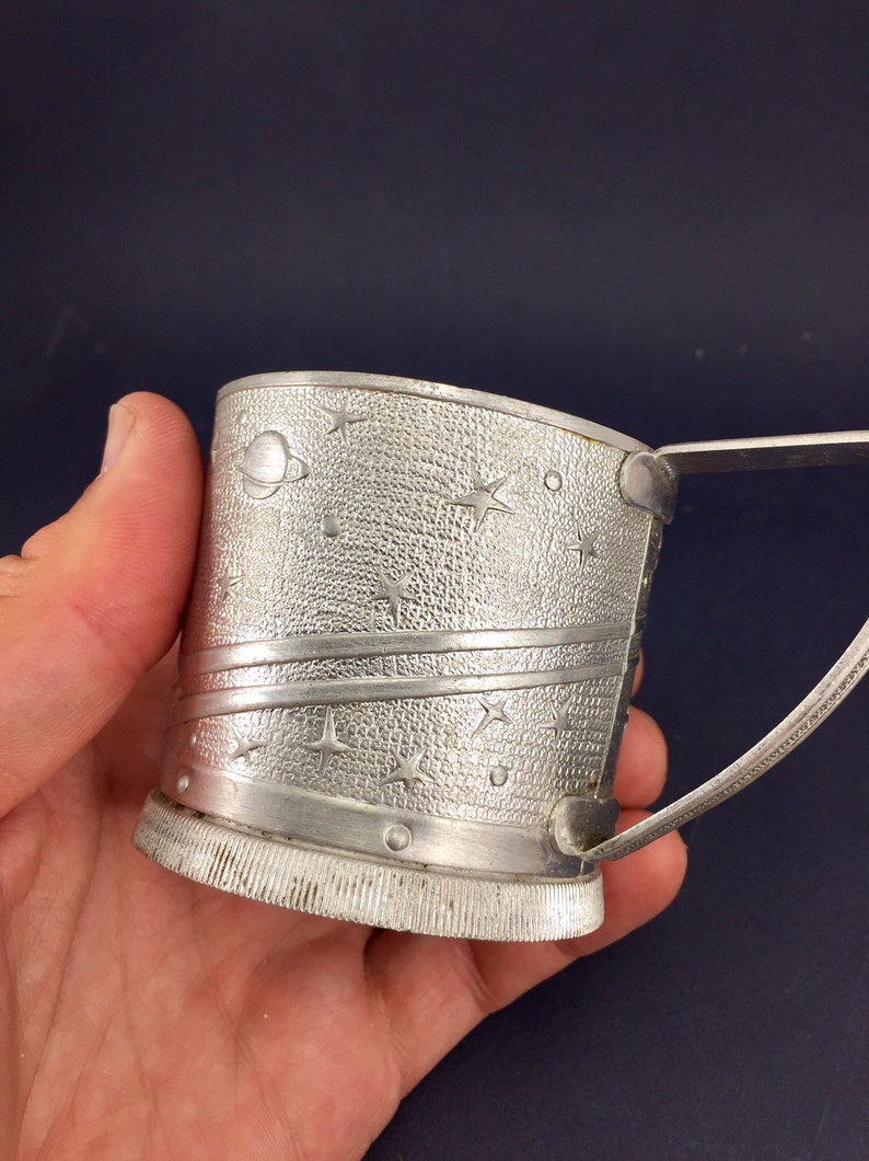 Peut inclure: Une tasse en m&eacute;tal argent&eacute; avec une poign&eacute;e, pr&eacute;sentant un motif sur le th&egrave;me de l'espace avec des &eacute;toiles et des plan&egrave;tes. La tasse a une surface textur&eacute;e et un fond c&ocirc;tel&eacute;.
