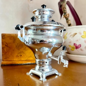 May include: A silver samovar with a black spout and handles. The samovar is on a wooden surface.