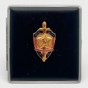 May include: A black cigarette case with a silver trim. The case features a gold emblem with a sword, star, and text in Cyrillic script. The emblem is set against a blue and red background.