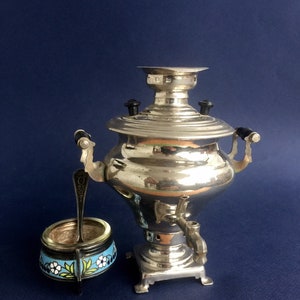 Puede incluir: Un samovar plateado brillante con asas negras y un grifo, junto a un peque&ntilde;o recipiente redondo con un dise&ntilde;o floral en azul, blanco y verde. Una cucharita est&aacute; en el recipiente. Sobre un fondo azul oscuro.