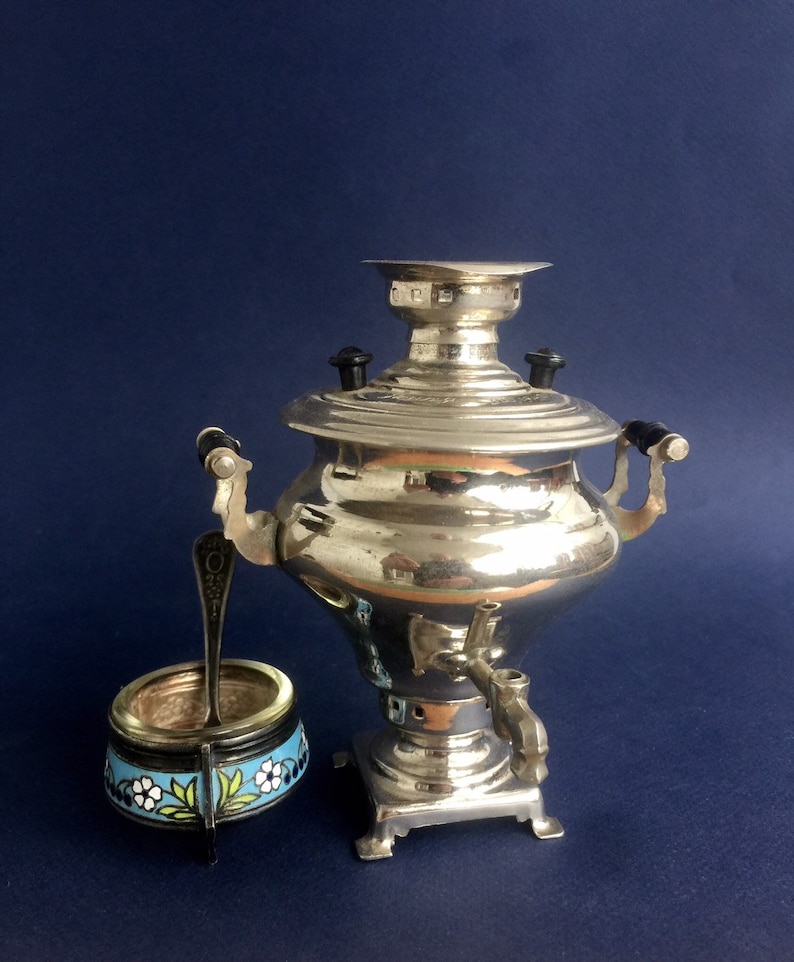 Puede incluir: Un samovar plateado brillante con asas negras y un grifo, junto a un peque&ntilde;o recipiente redondo con un dise&ntilde;o floral en azul, blanco y verde. Una cucharita est&aacute; en el recipiente. Sobre un fondo azul oscuro.