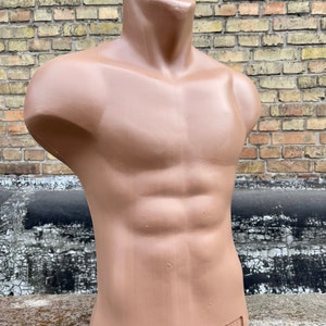 Puede incluir: Un maniquí de torso de plástico marrón con un pecho musculoso definido. El maniquí es sin cabeza y tiene un acabado liso y mate. La palabra "POWA" está impresa en la parte inferior del maniquí.