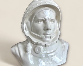 May include: White ceramic bust of a man wearing a space helmet. The bust is a realistic depiction of a cosmonaut.