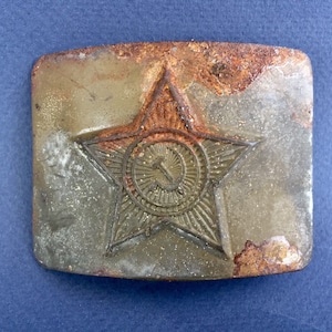 Pu&ograve; includere: Una fibbia per cintura in metallo arrugginito con una stella rossa a cinque punte e una falce e martello al suo interno. La stella ha un design testurizzato e in rilievo.