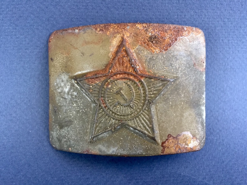 Pu&ograve; includere: Una fibbia per cintura in metallo arrugginito con una stella rossa a cinque punte e una falce e martello al suo interno. La stella ha un design testurizzato e in rilievo.