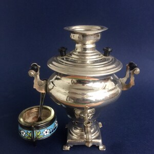 Puede incluir: Un samovar plateado con asas negras y un azucarero peque&ntilde;o a juego con una cuchara. El azucarero tiene una banda azul con dise&ntilde;os florales blancos y amarillos. El samovar es un dispositivo tradicional para preparar t&eacute;.