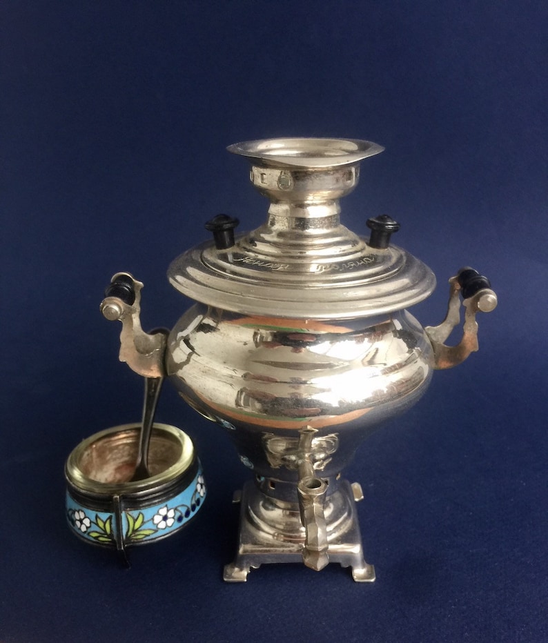 Puede incluir: Un samovar plateado con asas negras y un azucarero peque&ntilde;o a juego con una cuchara. El azucarero tiene una banda azul con dise&ntilde;os florales blancos y amarillos. El samovar es un dispositivo tradicional para preparar t&eacute;.