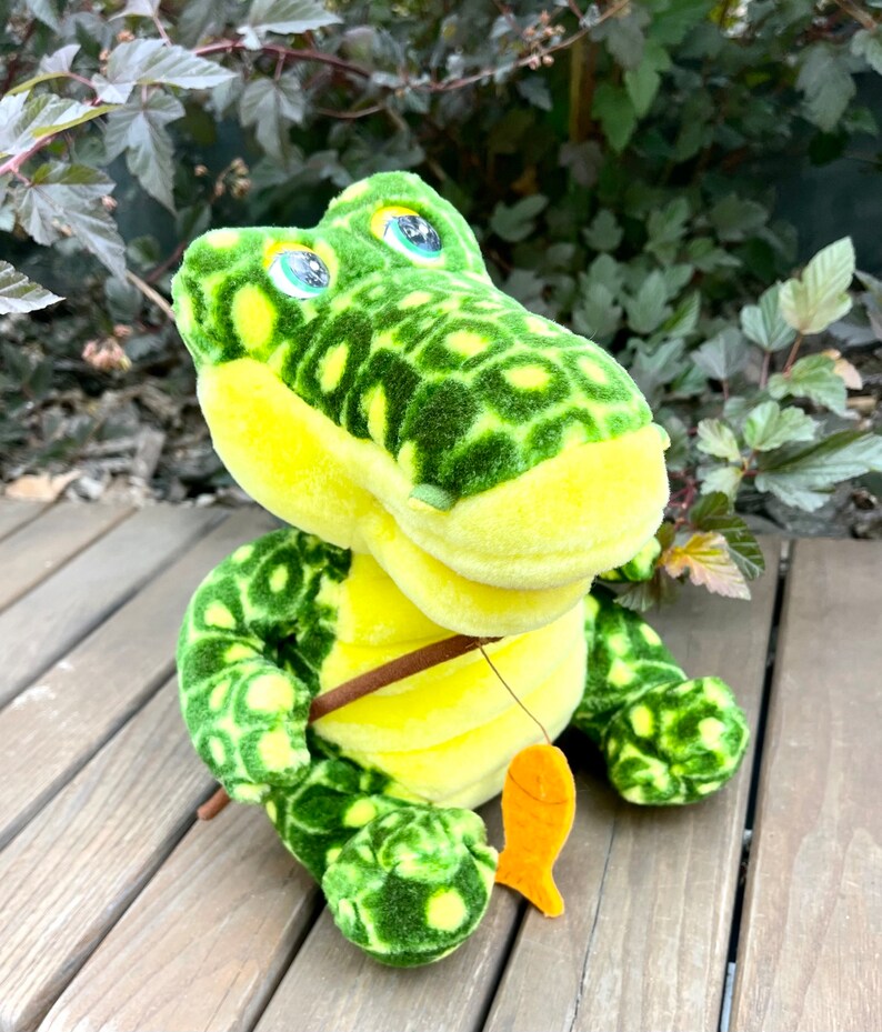 May include: A green and yellow plush crocodile toy with a fishing rod and a fish. The crocodile has a spotted pattern and big green eyes.