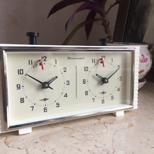 May include: A vintage chess clock with a white frame and two clock faces. Each face has black numbers, hands, and a red second hand. The clock has a black button on top and a silver frame around the clock faces.