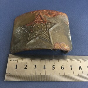 Pu&ograve; includere: Una fibbia per cintura in metallo verde rugginoso con una stella a cinque punte e un cerchio al centro. La stella ha un design in rilievo e il cerchio contiene una falce e un martello. La fibbia &egrave; larga circa 5 cm.