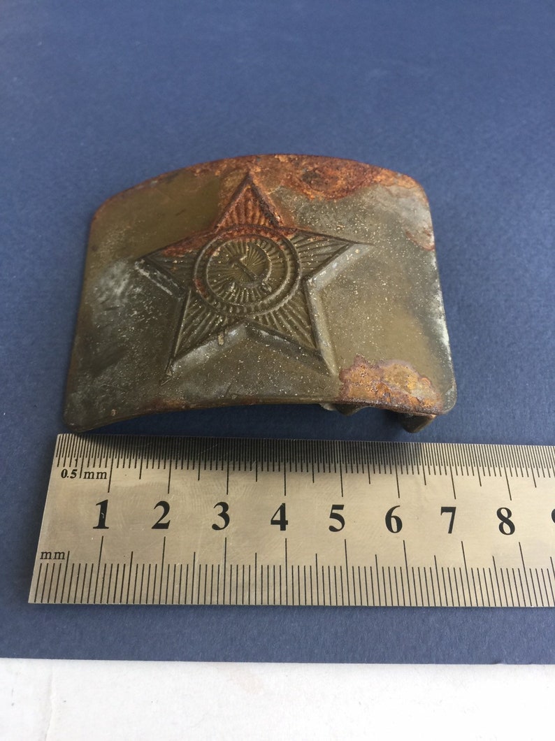 Pu&ograve; includere: Una fibbia per cintura in metallo verde rugginoso con una stella a cinque punte e un cerchio al centro. La stella ha un design in rilievo e il cerchio contiene una falce e un martello. La fibbia &egrave; larga circa 5 cm.