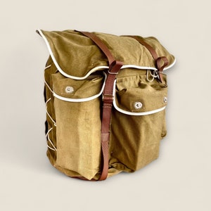 May include: A large, brown canvas backpack with white trim and brown leather straps. The backpack has two large pockets on the front and a smaller pocket on the top.