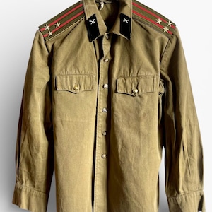 May include: Olive green military-style shirt with gold buttons and two chest pockets. The shoulders feature red and green stripes with gold stars. Black collar with white cross details. The shirt is hanging on a wooden hanger.
