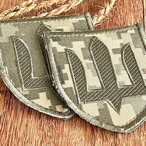 Pode incluir: Dois patches de tecido camuflado verde e castanho com símbolos de tridente ucraniano bordados a amarelo.