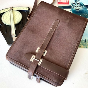 Könnte beinhalten: Eine braune Ledertasche mit einem Klappverschluss und einem Trageriemen. Die Tasche hat zwei silberne Schnallen und einen silbernen Verschluss. Die Tasche ist aus strukturiertem Leder.
