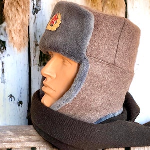Może przedstawiać: Brązowa i szara zimowa czapka z futerkiem i czerwonym emblematem gwiazdy. Czapka ma nauszniki, kt&oacute;re można zawiązać pod brodą.