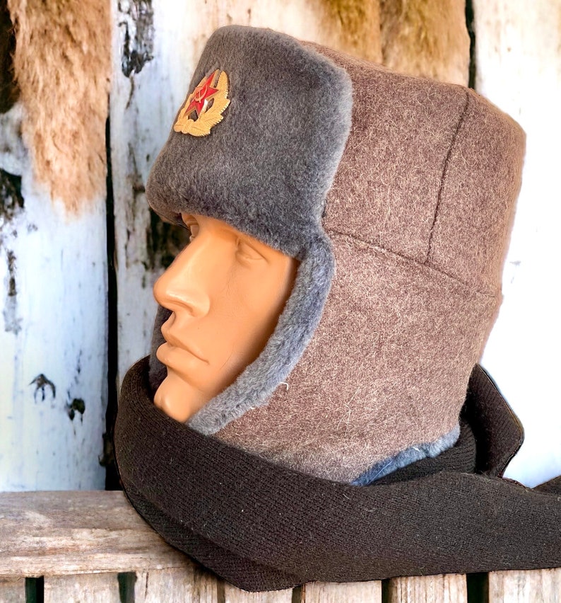 Może przedstawiać: Brązowa i szara zimowa czapka z futerkiem i czerwonym emblematem gwiazdy. Czapka ma nauszniki, kt&oacute;re można zawiązać pod brodą.