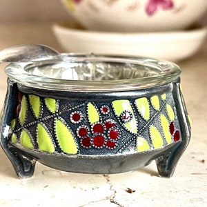 May include: A small, three-legged, glass and ceramic salt cellar with a green and red floral design.