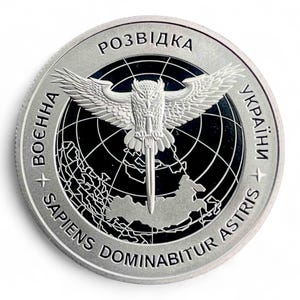 May include: A silver coin featuring an owl with outstretched wings, a sword, and a map. The coin has the words "Розвідка", "України", and "SAPIENS DOMINABITUR ASTRIS" around the edge. The coin is on a white background.