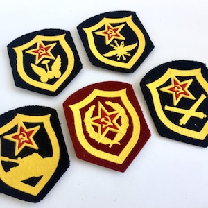 Puede incluir: Cinco parches bordados en forma de escudos con una estrella roja y otros símbolos en el centro. Los parches están sobre un fondo blanco.