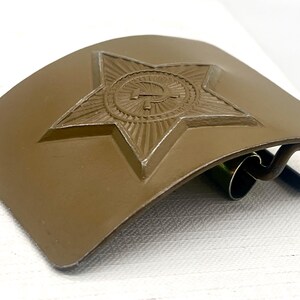 May include: A khaki-colored belt buckle with a raised star emblem featuring a hammer and sickle. The buckle has a curved shape and a metal clasp. The star is in the center of the buckle.