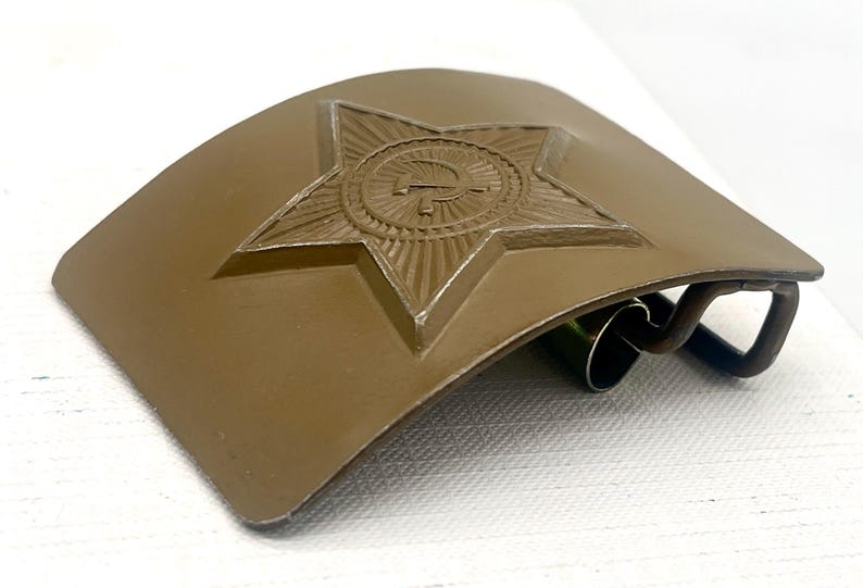May include: A khaki-colored belt buckle with a raised star emblem featuring a hammer and sickle. The buckle has a curved shape and a metal clasp. The star is in the center of the buckle.
