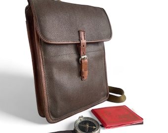 May include: A brown leather messenger bag with a brown leather strap and a silver buckle. The bag has a flap with a brown leather strap and a silver buckle. The bag is sitting on a white surface with a compass and a red booklet.