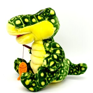 May include: A green and yellow plush dinosaur with a spotted pattern is holding a fishing rod with an orange fish attached. The dinosaur is sitting on a white background.