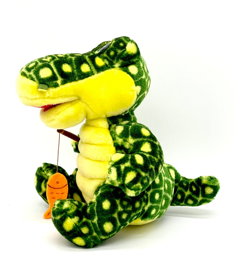 May include: A green and yellow plush dinosaur with a spotted pattern is holding a fishing rod with an orange fish attached. The dinosaur is sitting on a white background.