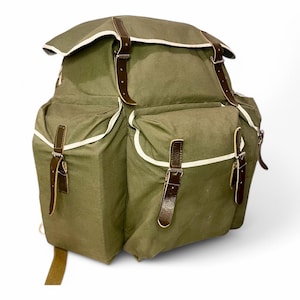 May include: Olive green canvas backpack with brown leather straps and silver buckles. The backpack has multiple compartments with white trim and a flap closure. A classic, durable design for outdoor adventures.