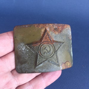 Pu&ograve; includere: Una fibbia per cintura in metallo arrugginito con una stella a cinque punte e una falce e martello all'interno di un cerchio. La stella &egrave; verde e il cerchio &egrave; dorato.