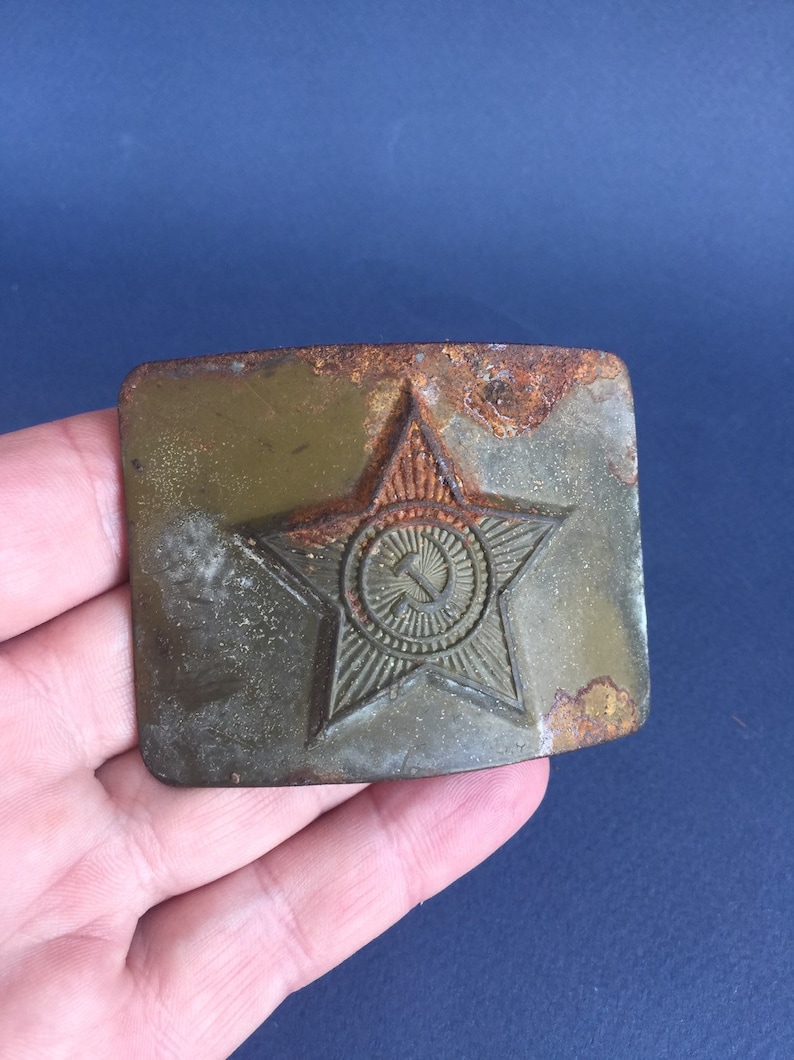 Pu&ograve; includere: Una fibbia per cintura in metallo arrugginito con una stella a cinque punte e una falce e martello all'interno di un cerchio. La stella &egrave; verde e il cerchio &egrave; dorato.
