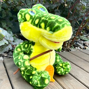May include: A plush green and yellow alligator toy holding a fishing rod with a small orange fish attached. The alligator has large blue eyes and a toothy grin.