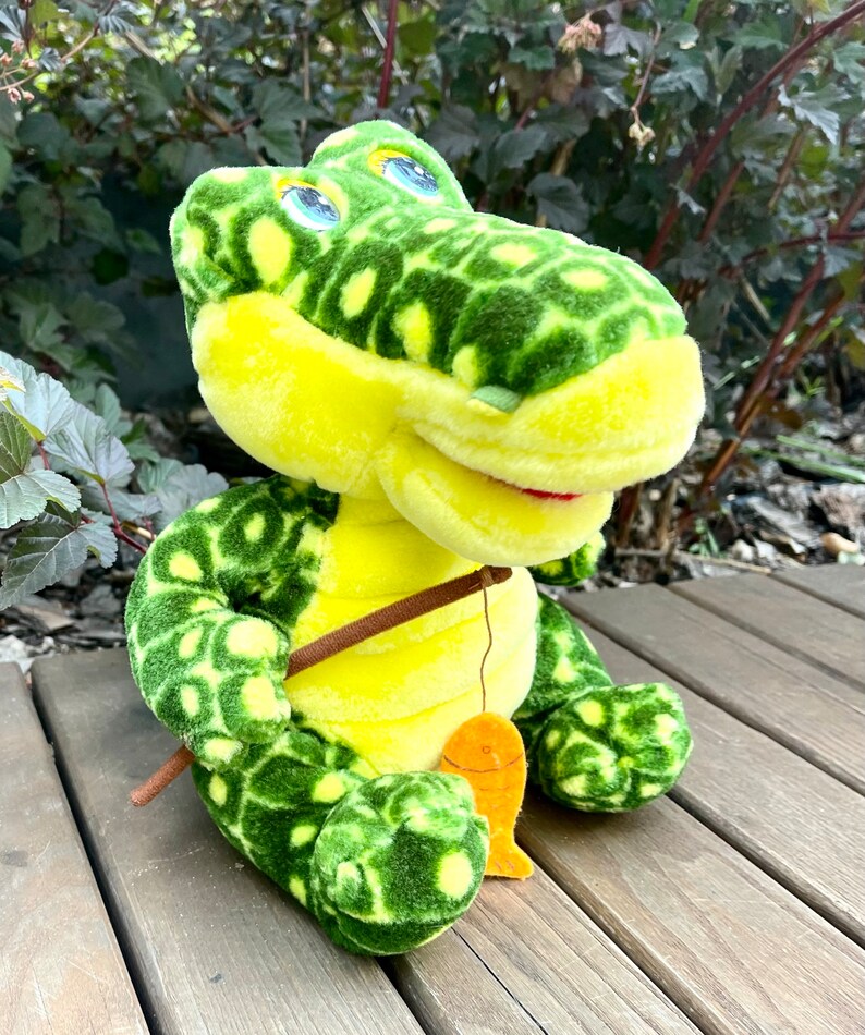 May include: A plush green and yellow alligator toy holding a fishing rod with a small orange fish attached. The alligator has large blue eyes and a toothy grin.