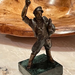 Peut inclure: Figurine en bronze d'un soldat en uniforme militaire, tenant un pistolet et une bouteille. La figurine est posée sur un socle en marbre vert. La statue a un design détaillé et est une pièce de collection vintage. Mesure environ 10 cm de haut.
