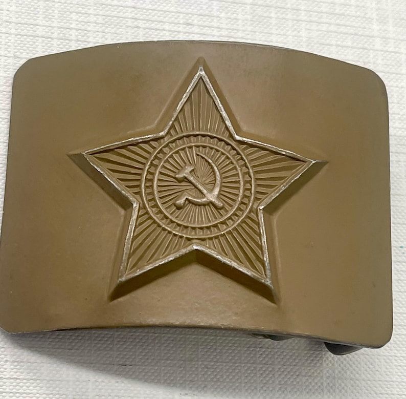 May include: A tan-colored belt buckle featuring a raised star emblem with a hammer and sickle symbol at the center. The star is surrounded by a sunburst design. The buckle has a rectangular shape.