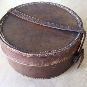 May include: A round, brown leather box with a stitched edge and a leather strap with a buckle. The box is slightly worn and has a vintage look.