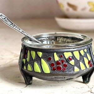 May include: A silver salt spoon rests on the rim of a decorative salt cellar. The cellar is made of metal and has a geometric design with yellow and red accents. The base of the cellar has three legs.