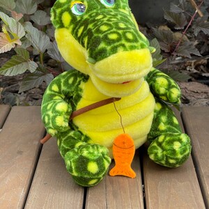 May include: A green and yellow plush crocodile toy holding a fishing rod with an orange fish attached. The crocodile has large, black eyes and a toothy grin.