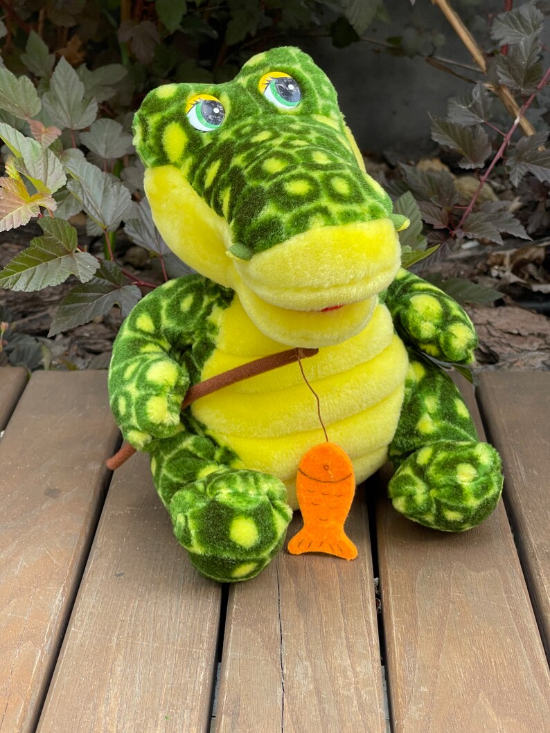 May include: A green and yellow plush crocodile toy holding a fishing rod with an orange fish attached. The crocodile has large, black eyes and a toothy grin.