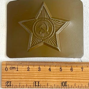 May include: A bronze-colored belt buckle featuring a raised star emblem with a hammer and sickle in the center. The buckle has a rectangular shape and is shown with a wooden ruler for scale.