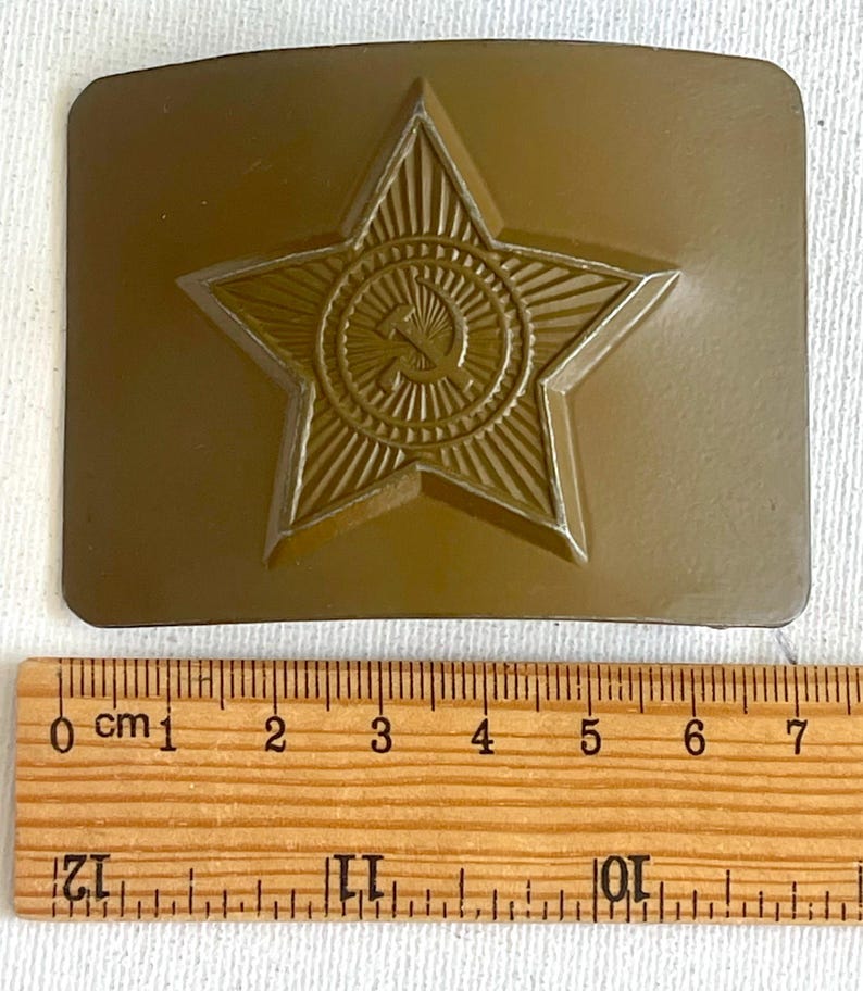 May include: A bronze-colored belt buckle featuring a raised star emblem with a hammer and sickle in the center. The buckle has a rectangular shape and is shown with a wooden ruler for scale.