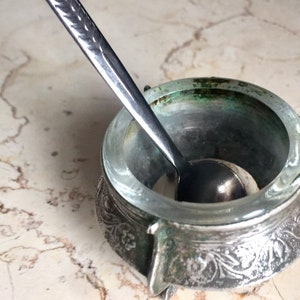 Könnte beinhalten: Eine silberne Salzdose mit einem verzierten Design und einem kleinen Löffel im Inneren. Die Salzdose hat einen klaren Glaseinsatz und steht auf einer Marmorfläche.