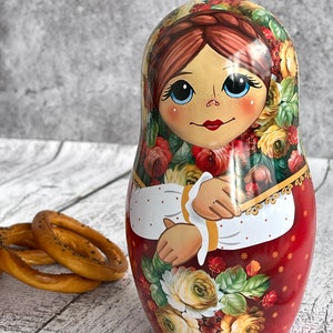 Op de afbeelding: Een traditionele Russische nestpop, of matrjosjka-pop, met een rood en wit bloemenpatroon. De pop is geschilderd met een glimlachend gezicht en draagt een rood en wit bloemenjurkje.