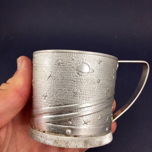 Peut inclure: Une tasse en m&eacute;tal argent&eacute; avec un motif sur le th&egrave;me de l'espace. La tasse a une poign&eacute;e et une surface textur&eacute;e avec des &eacute;toiles, des plan&egrave;tes et une com&egrave;te.