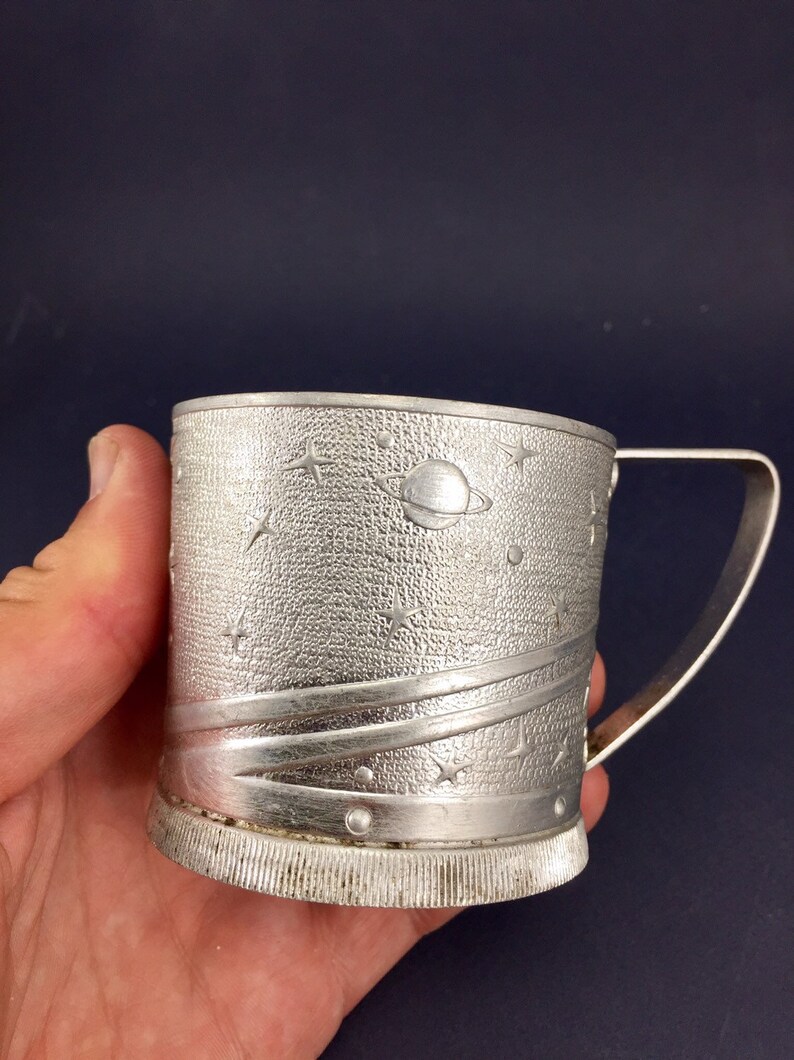 Peut inclure: Une tasse en m&eacute;tal argent&eacute; avec un motif sur le th&egrave;me de l'espace. La tasse a une poign&eacute;e et une surface textur&eacute;e avec des &eacute;toiles, des plan&egrave;tes et une com&egrave;te.
