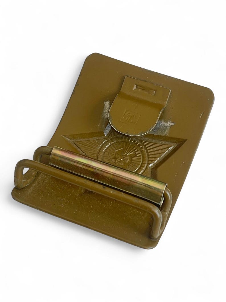 May include: Olive drab metal belt buckle with a star and wing emblem. The buckle has a rectangular base with a hinged clasp and a roller bar. The emblem features a star with wings and a circular design in the center.
