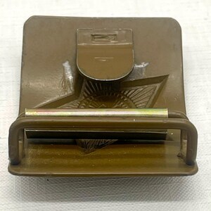 May include: A vintage olive drab metal belt buckle with a star emblem. The buckle features a rectangular base with a hinged clasp and a sliding bar for securing the belt. The design includes a detailed star and other embossed elements.