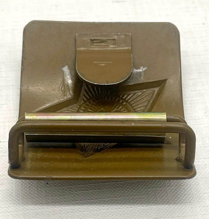 May include: A vintage olive drab metal belt buckle with a star emblem. The buckle features a rectangular base with a hinged clasp and a sliding bar for securing the belt. The design includes a detailed star and other embossed elements.